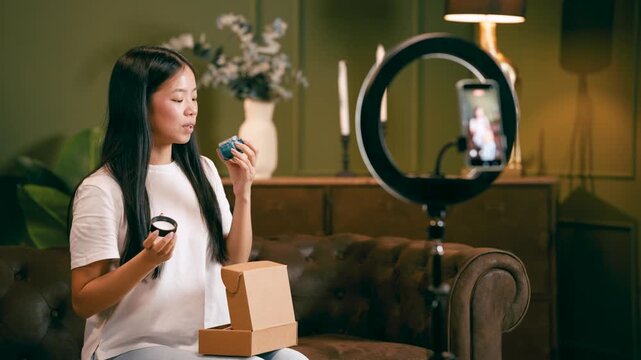 Young Asian influencer unboxing and reviewing a cosmetic cream on her social media channel, livestreaming with a smartphone and ring light