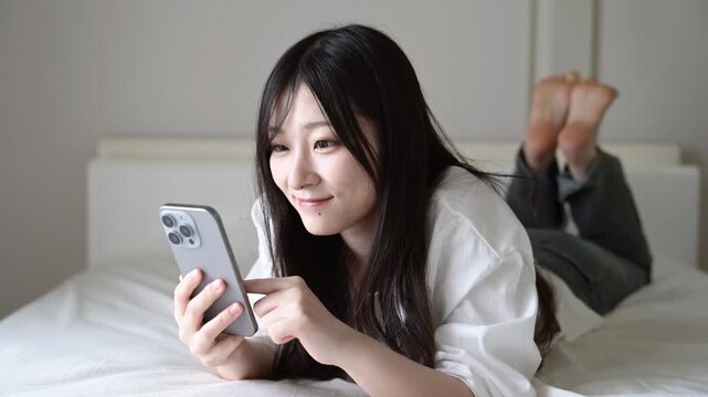 ベッドでスマートフォンを見ながら微笑む20代日本人女性