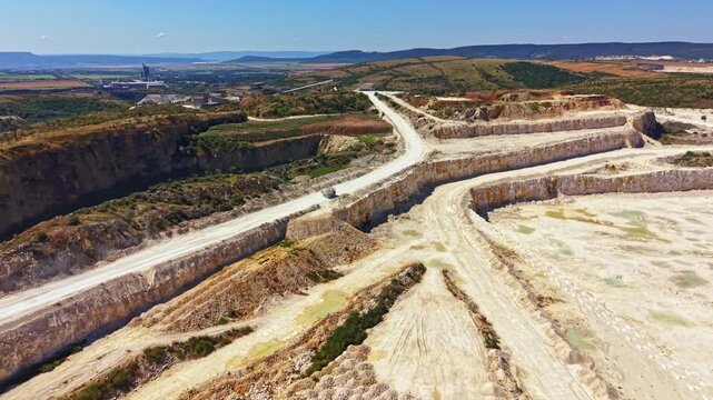 The scene shows a quarry with clear paths cut into the earth. Machines are at work moving materials. The sun shines down on open areas with hills visible in the distance.