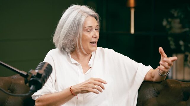 Elegant senior woman with gray hair actively gesturing and speaking into a microphone, recording a podcast or radio show in a studio