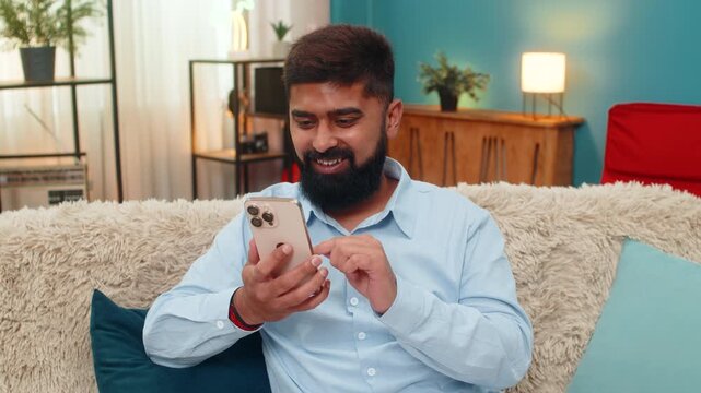 Indian man at home lifts smartphone camera smiles takes selfie captures joyful moment shares soon. Arabian guy on sofa posts new photo to social media feeling fun connection online presence happiness.