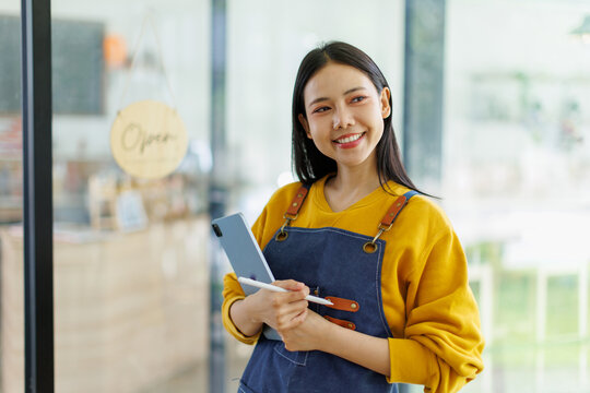 Woman small business owner in apron smiling holding tablet and stylus at coffee shop entrance with open sign