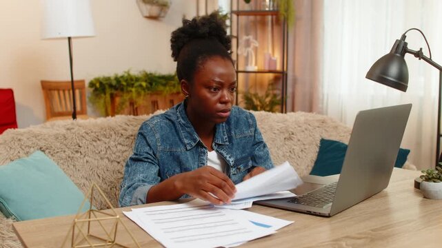 African American young woman sitting at table on home sofa nervously checks utility bills using laptop, printed papers. Black girl looks stressed overwhelmed trying to manage household payments online