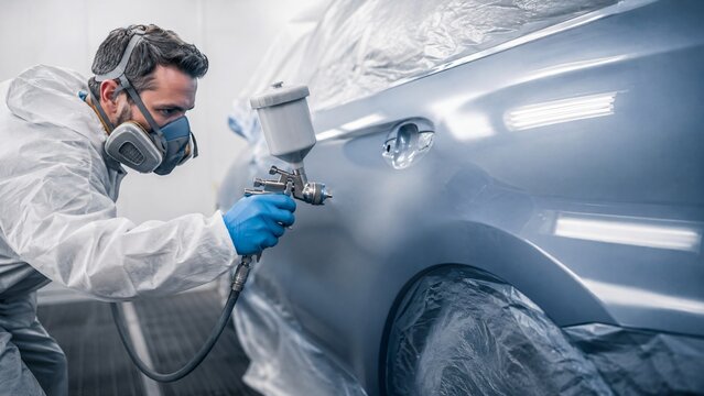 Painter using spray gun to paint car body in garage. Professional auto repairman in protective suit and respirator. Automotive bodywork service and vehicle restoration process.