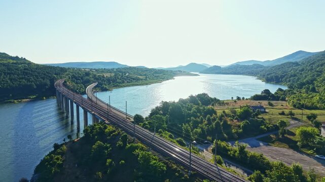 A river flows beside a bridge that supports a train track. Green trees line the banks, and mountains rise in the background. The sun shines brightly over the landscape.
