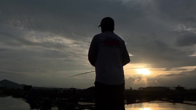 Low-angle shot captures a gray and orange sunset over the fish ponds of Cirata Reservoir. A fisherman casts a line from a boat surrounded by aquaculture cages as the sun nears the horizon. 