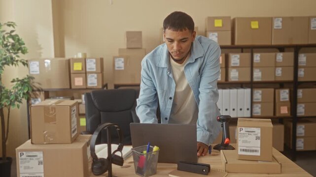 Man leaning on desk with laptop and barcode scanner amid stacked shipping boxes in a small business building; ecommerce confidence.