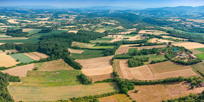 Panoramic aerial view of a patchwork countryside with hay bales, scattered farmhouses, and wooded hills under a summer sky.