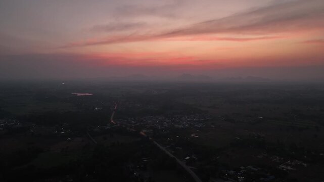 Aerial footage rural village in Nong Bua Lamphu nestled in nature under a vibrant orange sunset sky. Street lights and houses begin to illuminate as dusk transitions into night.