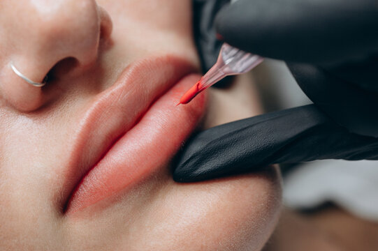 Macro close up of a permanent makeup artist performing a lip blush tattoo procedure with a PMU machine in a professional beauty salon.