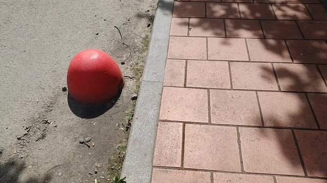 Red bollard on sidewalk with shadows and greenery in background 