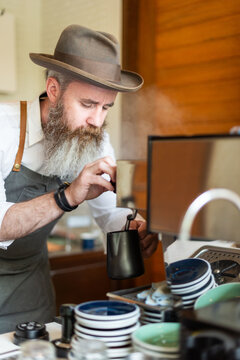Bearded man in hat making coffee at a cafe with focused expression.