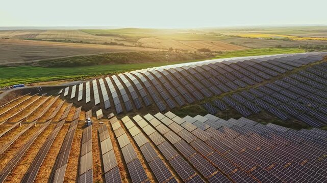 Workers maintain solar panels in a large field as the sun sets. Rows of panels stretch across the ground creating a grid. Green fields and hills are visible in the distance.