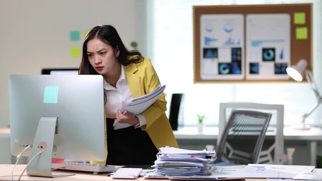Businesswoman experiencing stress working at computer. Young businesswoman hold document while focusing intently on a computer in office, showcasing the challenges of work-related stress and pressure.