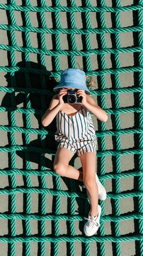 Young child discovering photography, looking through a vintage camera lens while relaxing on a vibrant green rope net outdoors