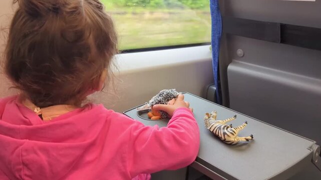  girl in a pink sweater plays with toy animals on a folding table during a train ride. Concept of family travel, kids' entertainment during a journey, and childhood memories.