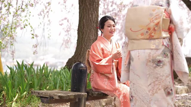Soft pink blossoms frame a tranquil garden scene. Woman in elegant kimono walks gently beneath cherry branches. She pauses near a stone basin, perhaps to cleanse her hands. Another woman sits nearby
