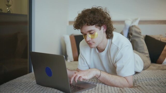 Young man lies on bed using laptop while wearing under-eye patches for skincare in a modern room with simple decor