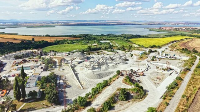 Large granite quarry shows stone piles, machinery, and workers. Nearby, fields and a lake are visible under a clear sky. Trees line the edges of the area.