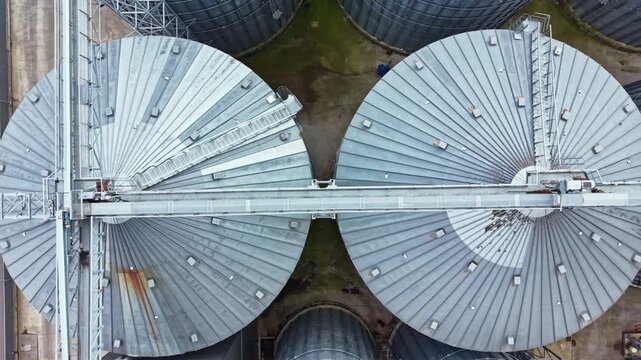 An aerial view shows several large, circular grain storage silos in a farming location. The silos have metal roofs and stand close together on flat ground.