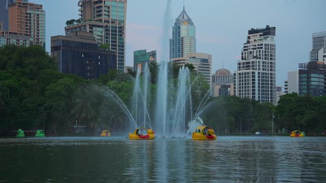 Lumpini Park fountain and pedal boats on lake with Bangkok city skyline, People riding duck boats on water among skyscrapers in Thailand, slow motion 4k