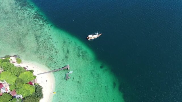 High-angle drone shot captures a traditional pinisi boat sailing on turquoise Sulawesi waters. The vessel nears a tropical island with white sand and lush greenery, creating a vibrant color contrast. 