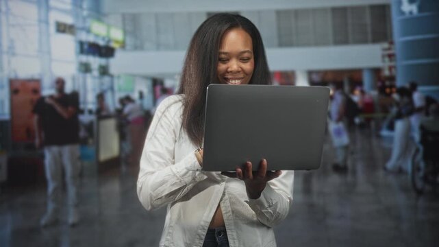 Woman holding laptop with fingers on keyboard and gaze fixed on screen in crowded airport terminal near checkin kiosks, wearing white shirt and jeans; focused productivity.