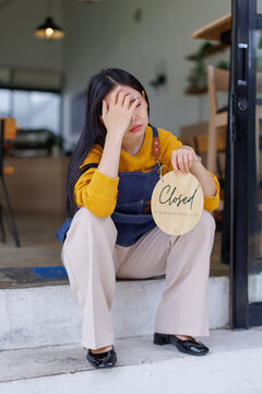 Young female shop owner feeling disappointed, sitting on steps with her head in hands, indicating a store closure