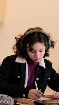 Focused teenage student in casual jacket sits at table with laptop and notebook, studies with headphones on to block noise, concept concentration and productivity for teens.
