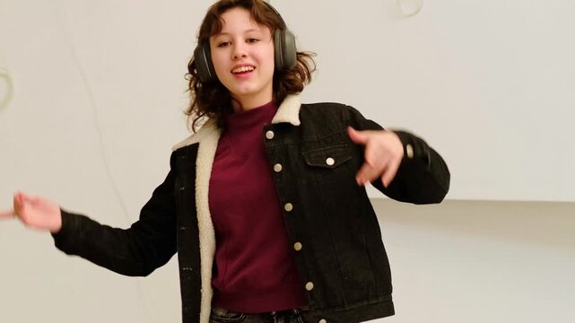 Teen girl in casual jacket listens to favorite playlist in wireless headphones and dances playfully, concept positive emotions and stress relief for teenagers.