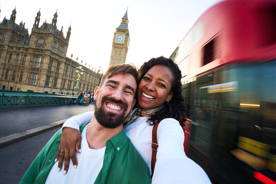 A cheerful men and women hold the camera for a selfie on westminster bridge. They are smiling with the iconic clock tower and a moving red double decker bus in the background.