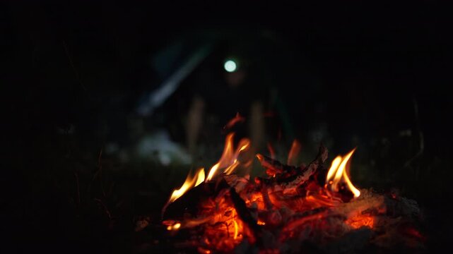 Warm campfire with glowing embers and stacked firewood burning beside a camping tent, illustrating outdoor camping, survival skills, nature adventure, and peaceful night recreation.