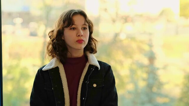 Teenage girl in a warm jacket turns her head and looks around by a large window as if waiting for someone who has not arrived, her face sad and uncertain. This moment reflects the loneliness and