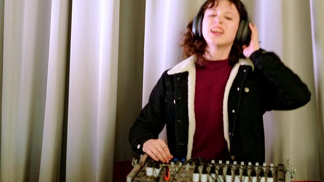 Young girl in casual jacket works carefully at a mixing console with headphones on, creating modern electronic music and developing creative technical skills.