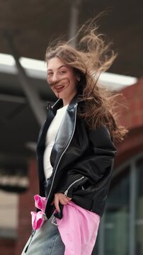 Young woman in leather jacket turns with her hair blowing in the wind, takes out her phone and starts reading a new message on a modern city street.