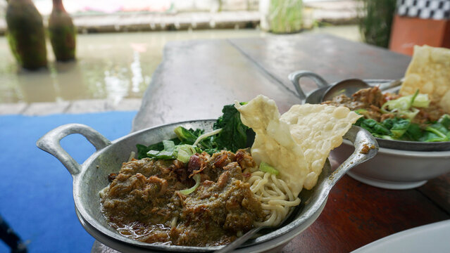 Indonesian chicken noodle mie ayam topped with savory minced chicken and mustard greens served in a metal bowl on a wooden table near a koi fish pond.