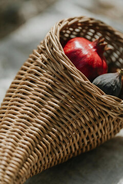 Detailed close-up of a woven wicker cornucopia horn filled with red pomegranate and dark fig in warm sunlight. Thanksgiving autumn harvest produce concept.