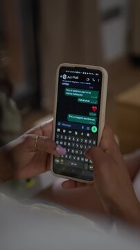 Vertical. Black Latina millennial woman lying comfortably at home while typing messages on her smartphone. Over-the-shoulder view showing digital communication, relaxation and everyday mobile use.