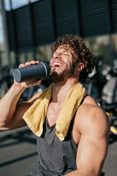 Smiling muscular man laughing while drinking protein shake at gym with yellow towel for fitness poster and lifestyle advertisement