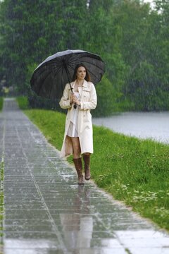 Woman walks on stone path next to body of water during rain. She holds black umbrella while rain falls around her. Green trees surround area creating natural setting