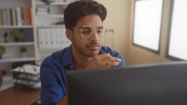 Man with round glasses holds a red pen to his lips while studying a laptop at a desk with bookshelf and file trays in a building; thoughtful focus.