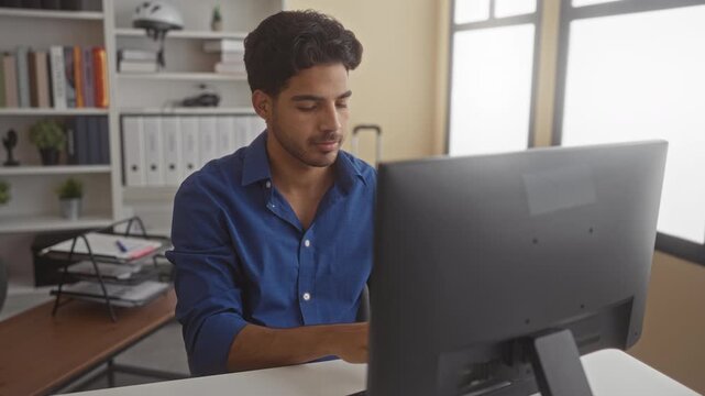 Man in blue shirt stretches and raises both hands above shoulders while smiling at a desktop monitor, holding a pen on the desk in building; relaxed satisfaction.