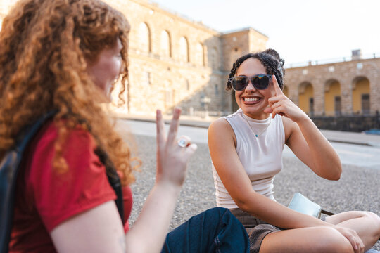 Two diverse young female friends sit on the pavement of a historic city square, laughing and talking. They use expressive hand gestures during their conversation.