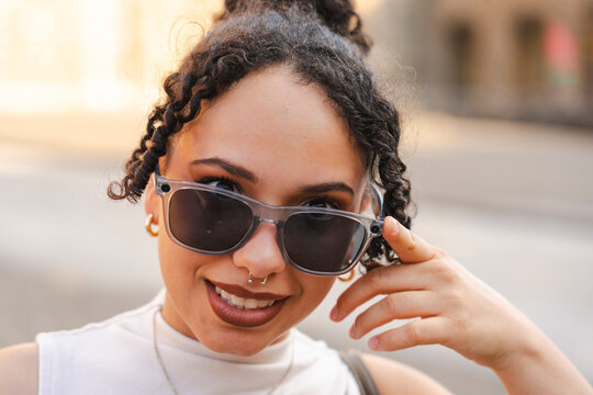 A stylish young woman with curly hair smiles cheerfully while peering over her sunglasses on a city street. This close-up portrait captures her confident, trendy look.