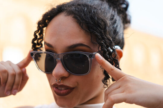 A close-up portrait of a stylish young woman with curly dark hair and a septum piercing, adjusting her fashionable grey sunglasses while posing outdoors in sunlight.