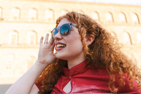 A cheerful young woman with long curly red hair and sunglasses smiles as she holds a hand to her mouth, calling out to a friend near a historic stone building.