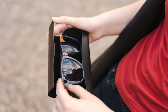 A woman wearing a red top holds a black leather case containing stylish grey sunglasses, with a reflection of the blue sky and clouds visible on the lenses.