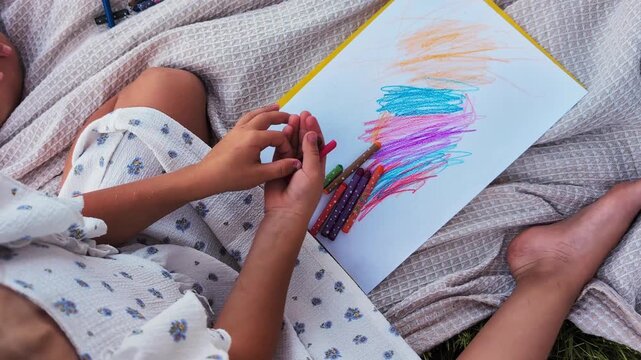 A child sits on a soft blanket in a grassy area, using crayons to create colorful drawings on a white sheet of paper