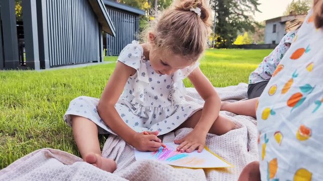 A young girl is focused on coloring with crayons while sitting on a blanket. She is in a grassy area with trees and buildings in the background