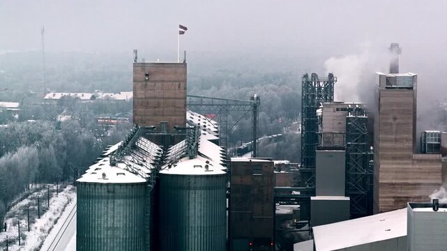 Aerial view of industrial silos and factory in winter with snow-covered trees and railway tracks in Latvia.